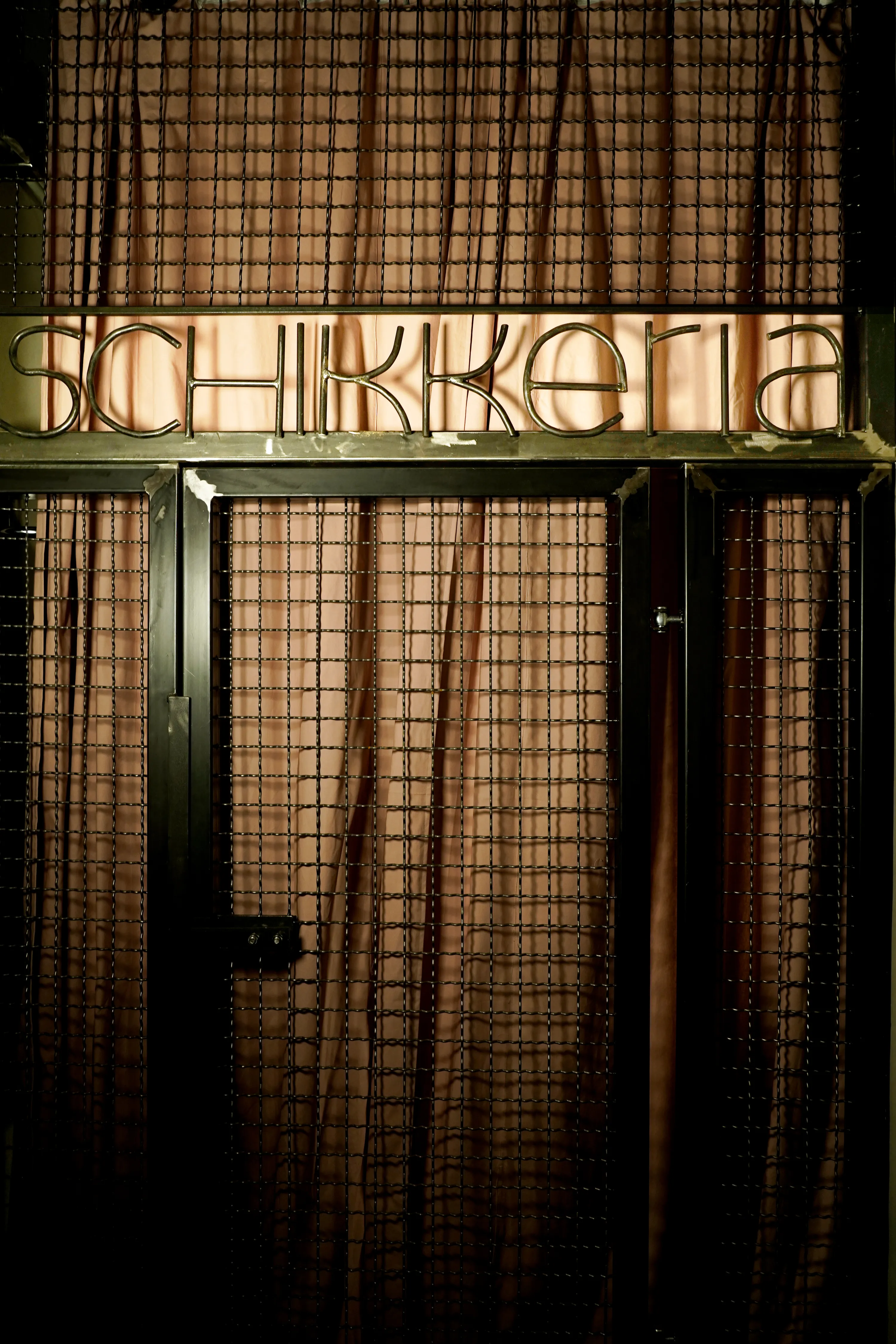 Handgefertigtes Eingangstor aus Metall und Drahtgeflecht mit dem Schriftzug „SCHIKKERIA“ im Wimpern- und Sugaringstudio Schikkeria in Leipzig. Dahinter hängt ein altrosa Vorhang.